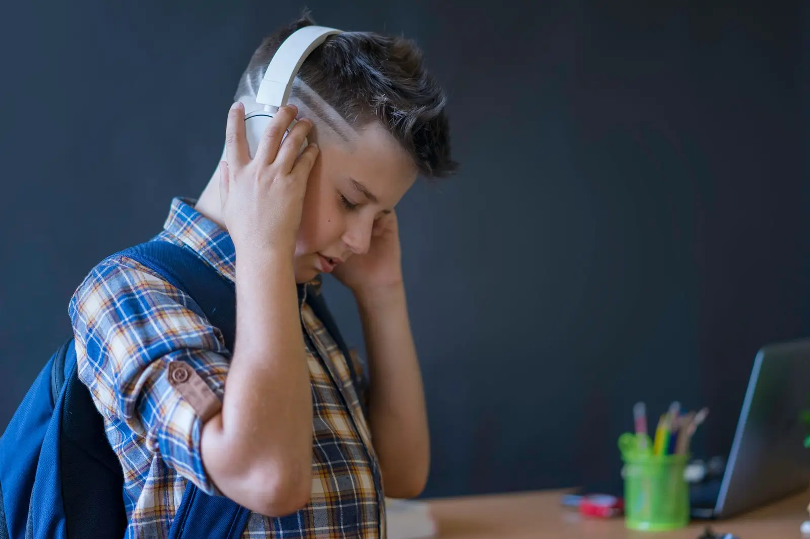 A boy with a backpack holding headphones over his ears at a desk