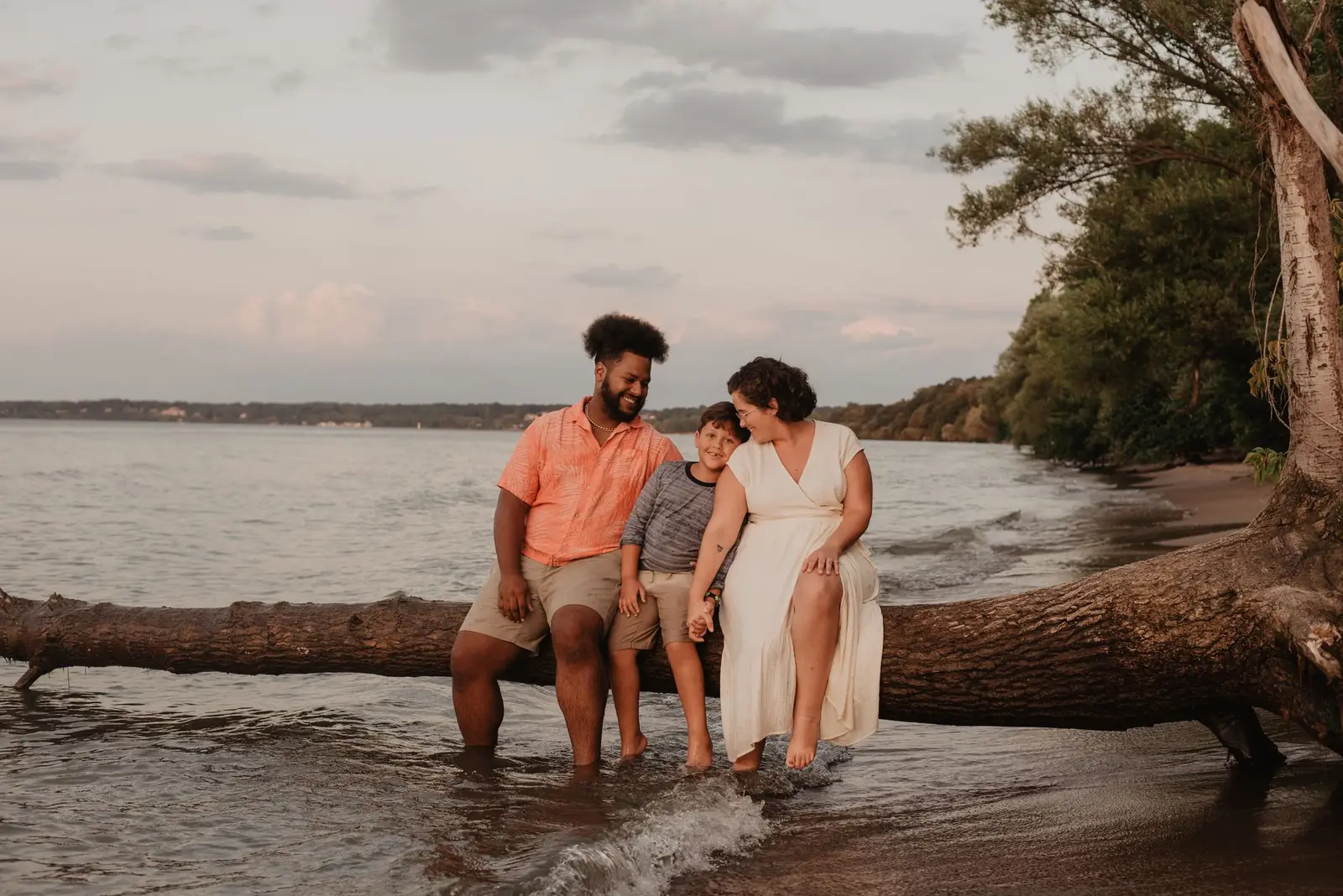 A family of three sitting together on a fallen tree by the water