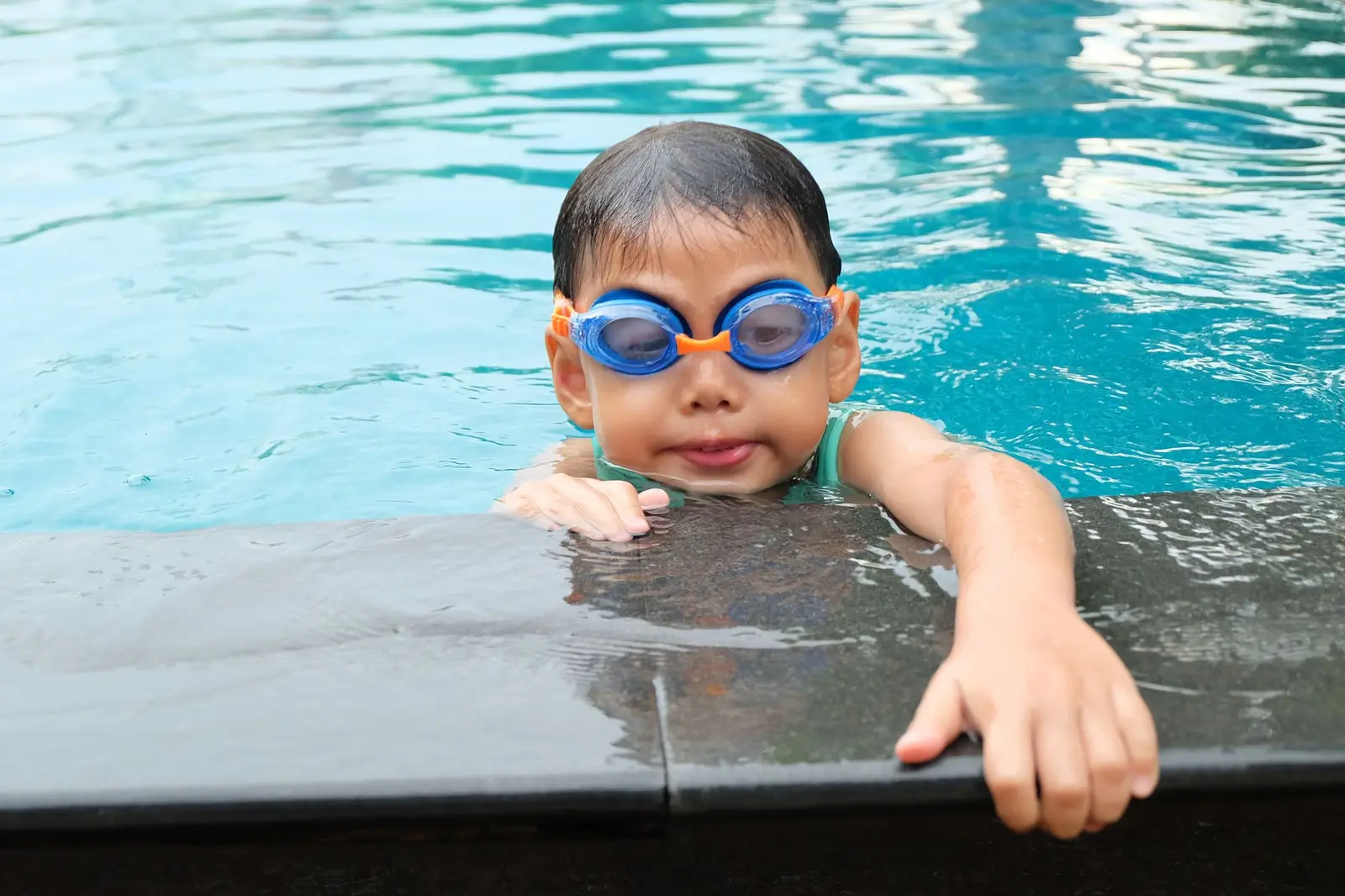 A young child in blue goggles resting at the edge of a pool