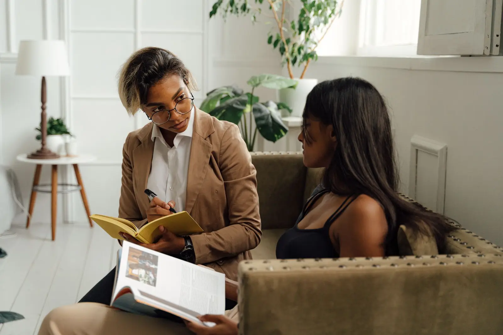 Two women talking in a bright room, one taking notes in a notebook