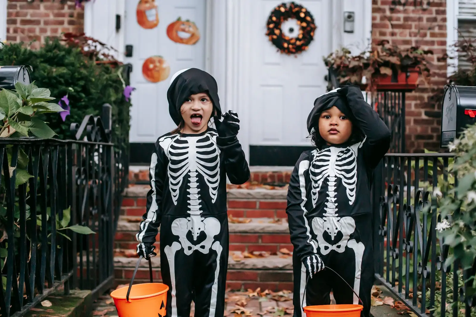 Two children in skeleton costumes holding orange buckets outside a house