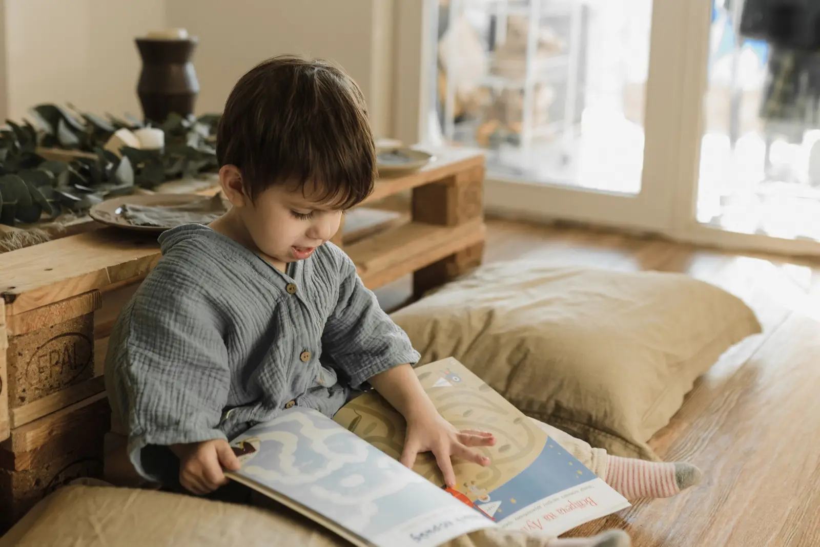 A young child reading a book on floor cushions by a window