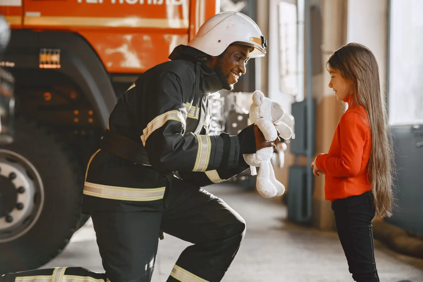 A firefighter kneeling to hand a soft toy to a young girl
