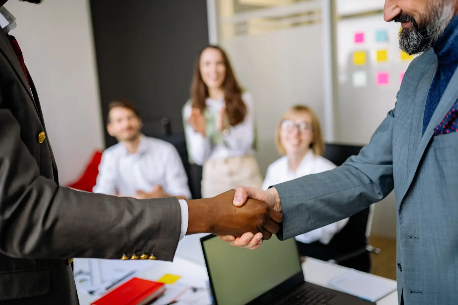 Two people shaking hands in an office with colleagues in the background