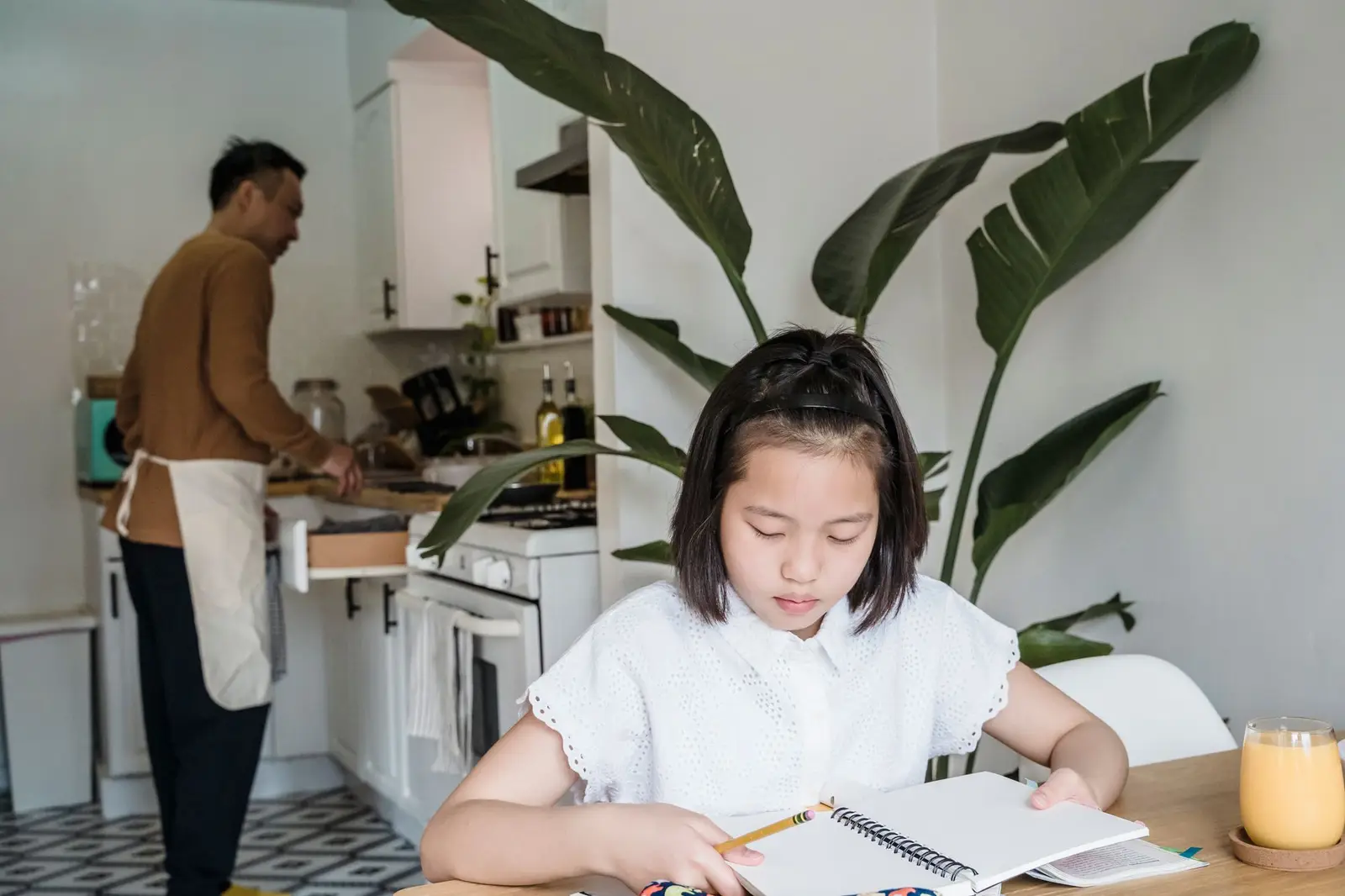 A girl doing homework at the kitchen table while her father cooks