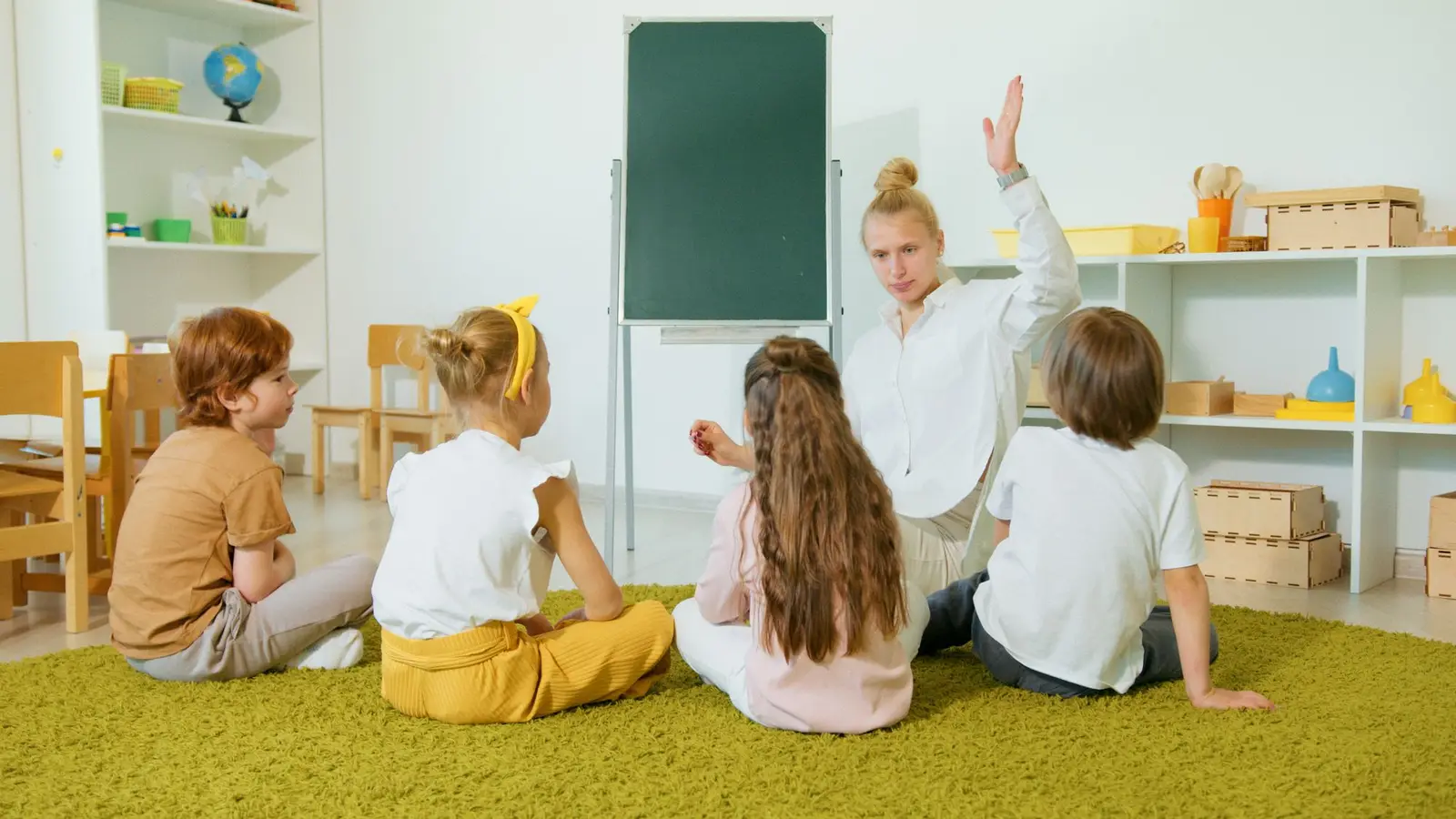 A preschool teacher with her hand raised talking with children sitting on the floor
