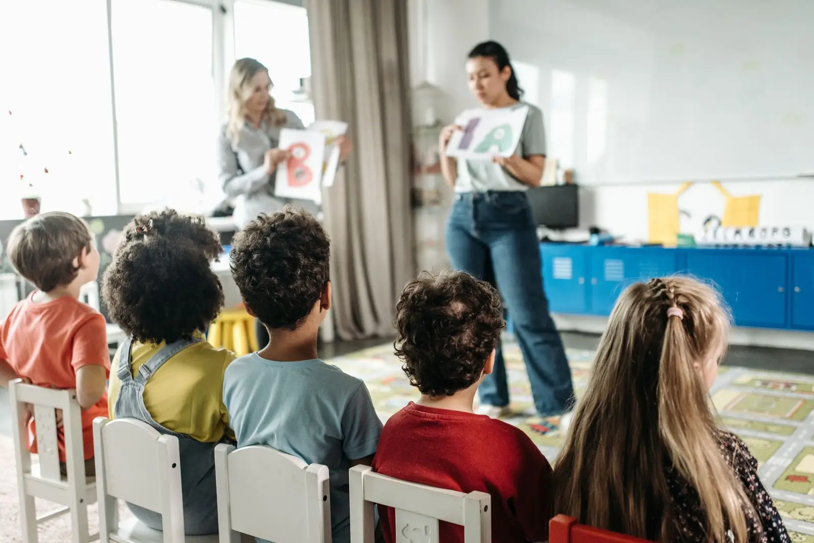 Children sitting on small chairs watching teachers hold up picture cards