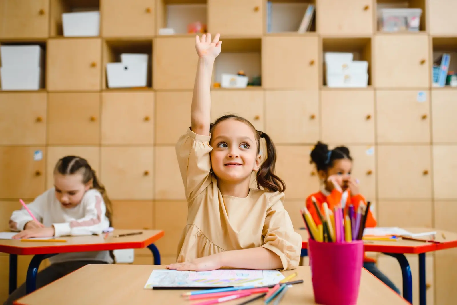A smiling girl raising her hand in a classroom with two classmates working nearby