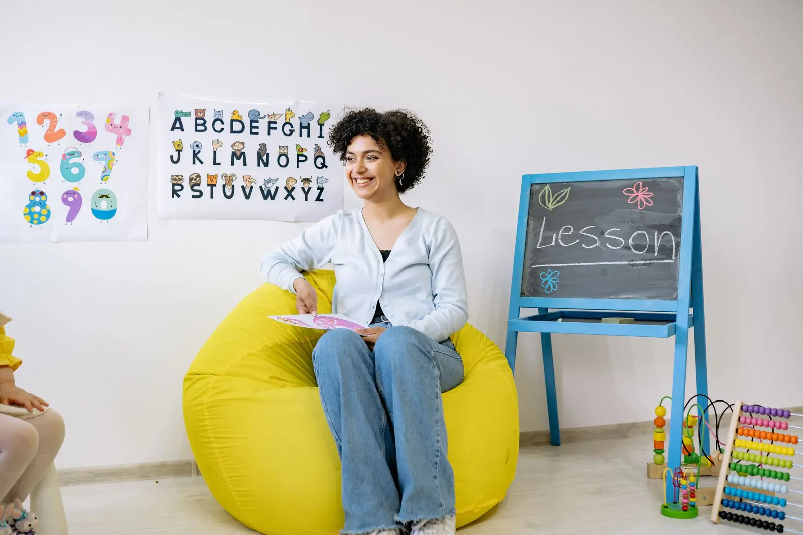 A smiling educator sitting on a beanbag holding a book in a preschool classroom