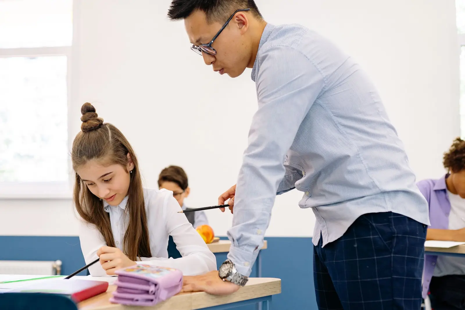 A teacher leaning over to help a student with her work at a classroom desk