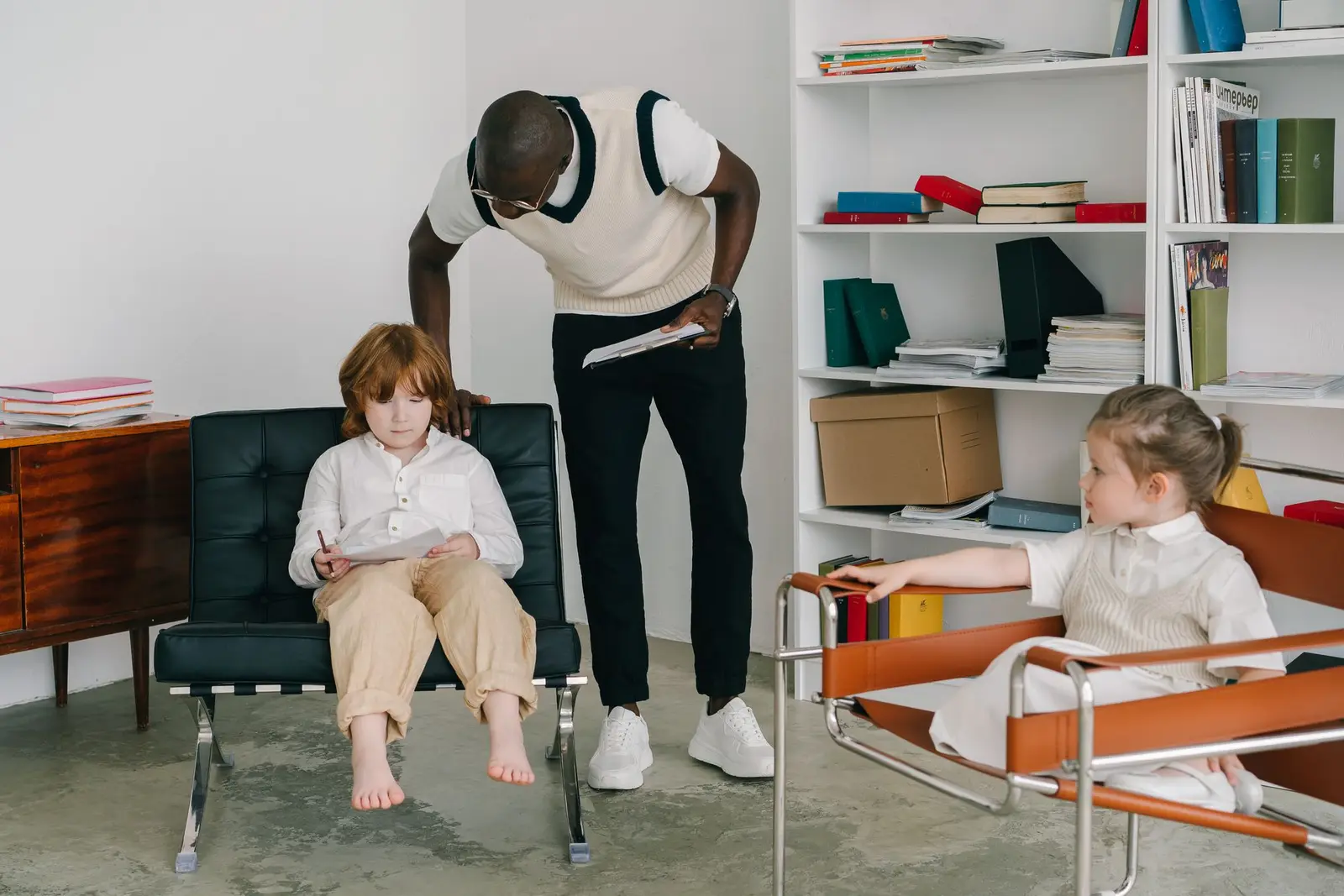 A therapist bending to talk with two children sitting in chairs in a bright room