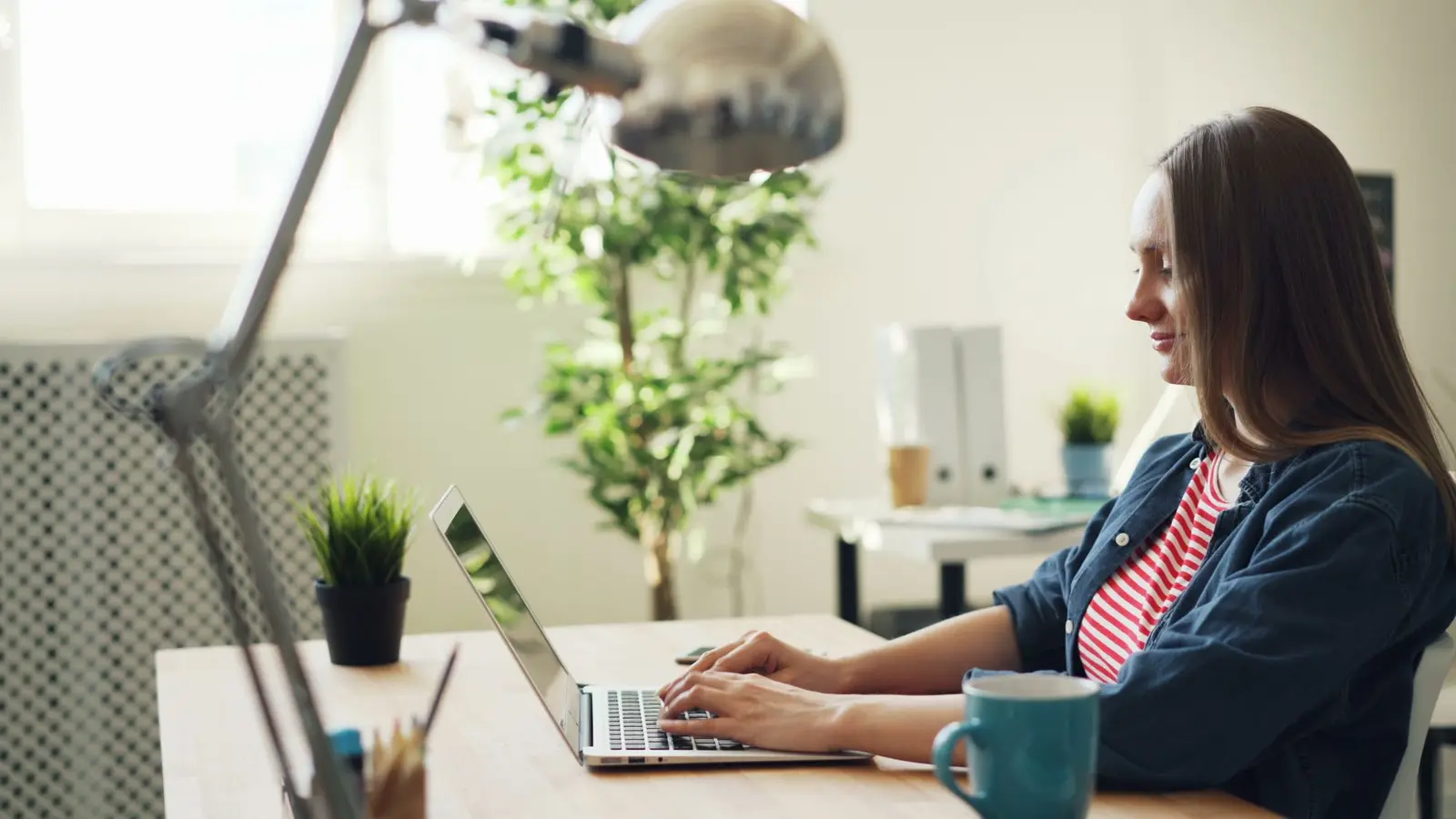 A woman smiling while working on a laptop in a bright office with plants