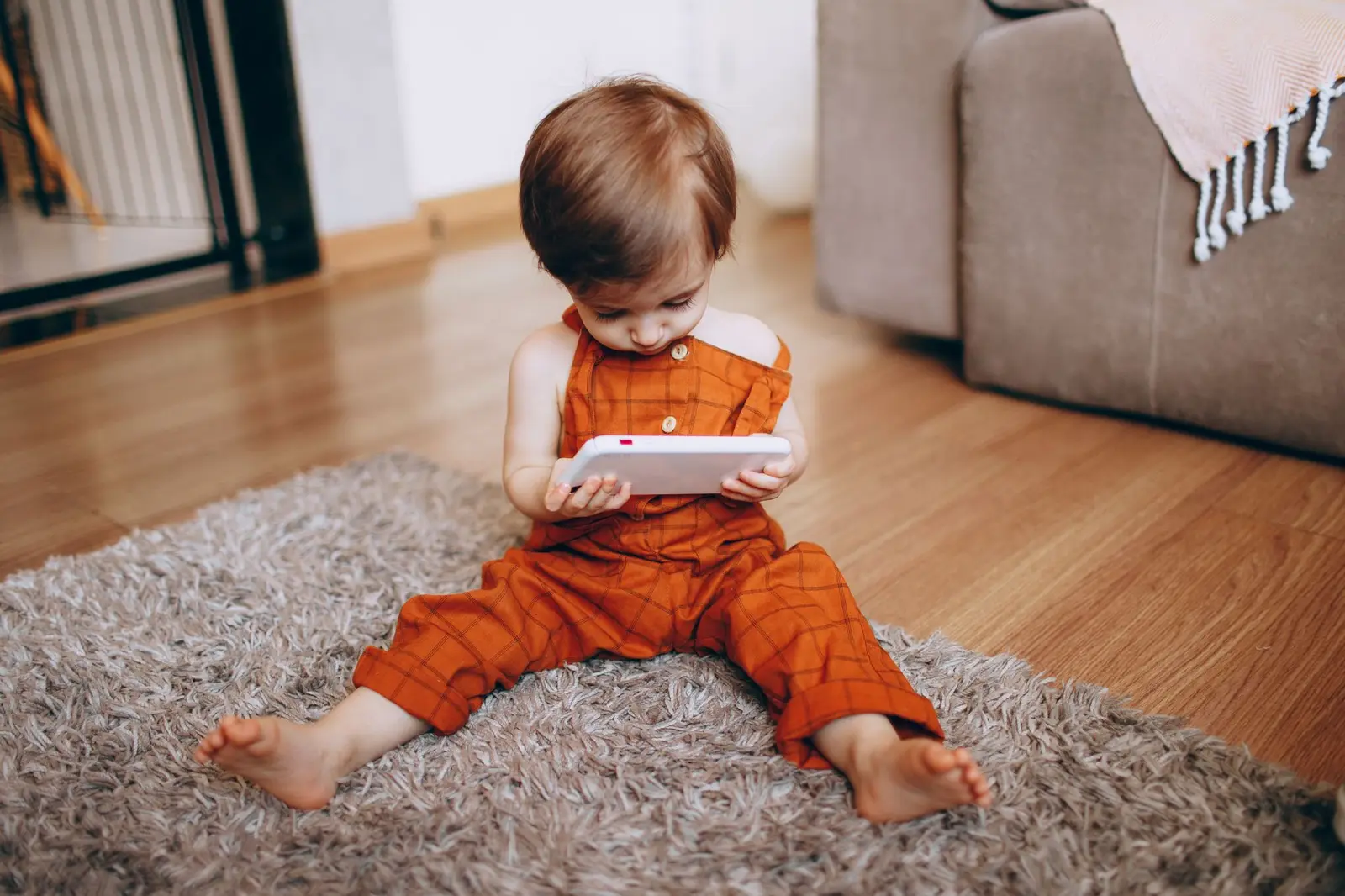 A toddler in orange overalls sitting on a rug looking at a tablet