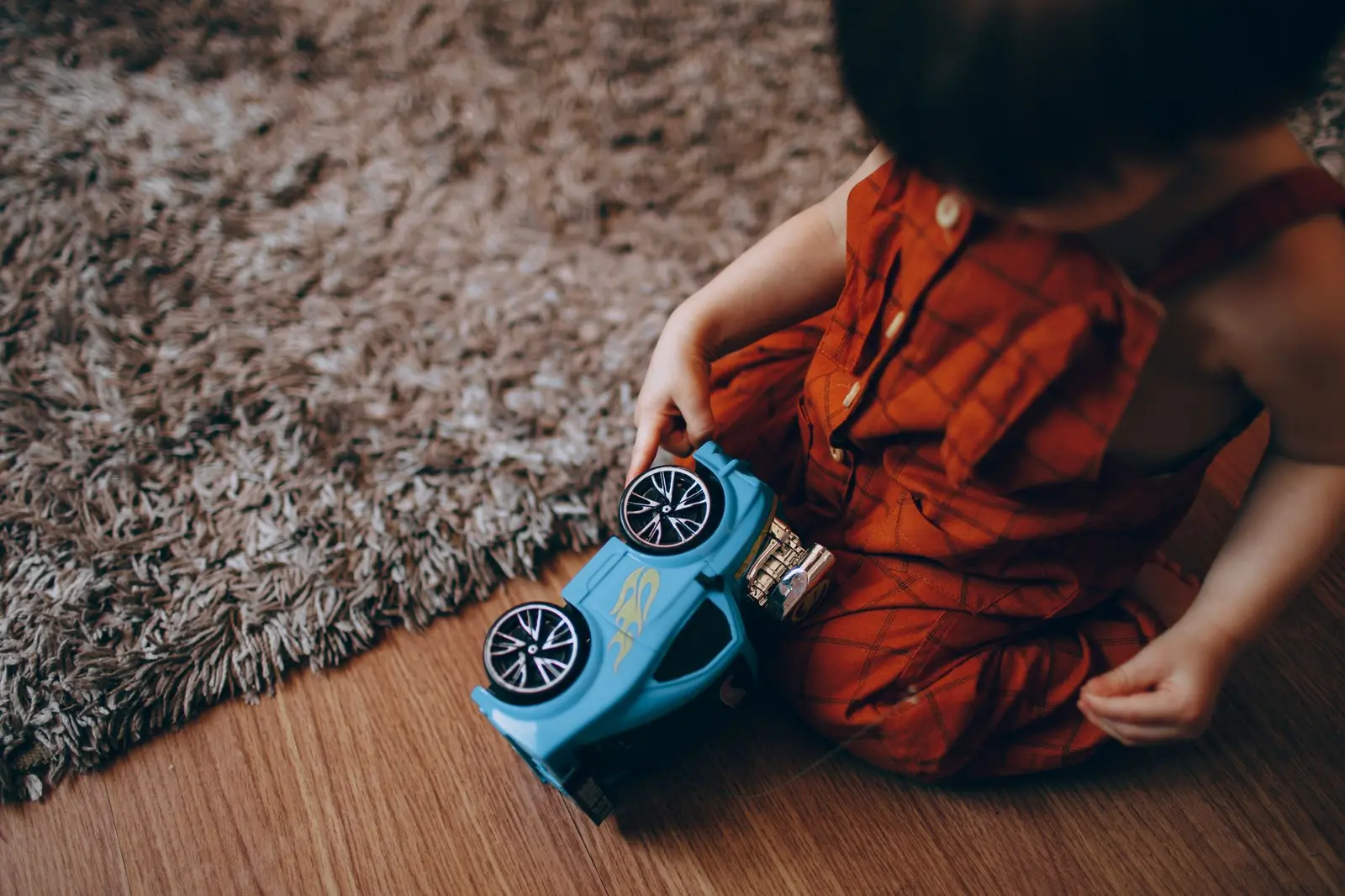 A toddler in red overalls playing with a blue toy car on a rug