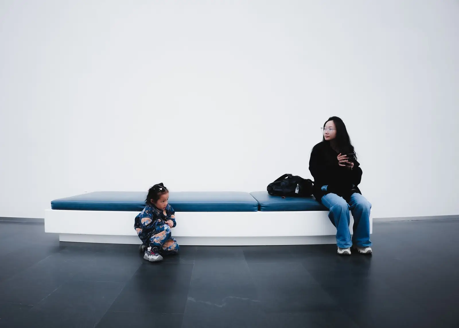 A small child crouching near a bench while a woman waits nearby