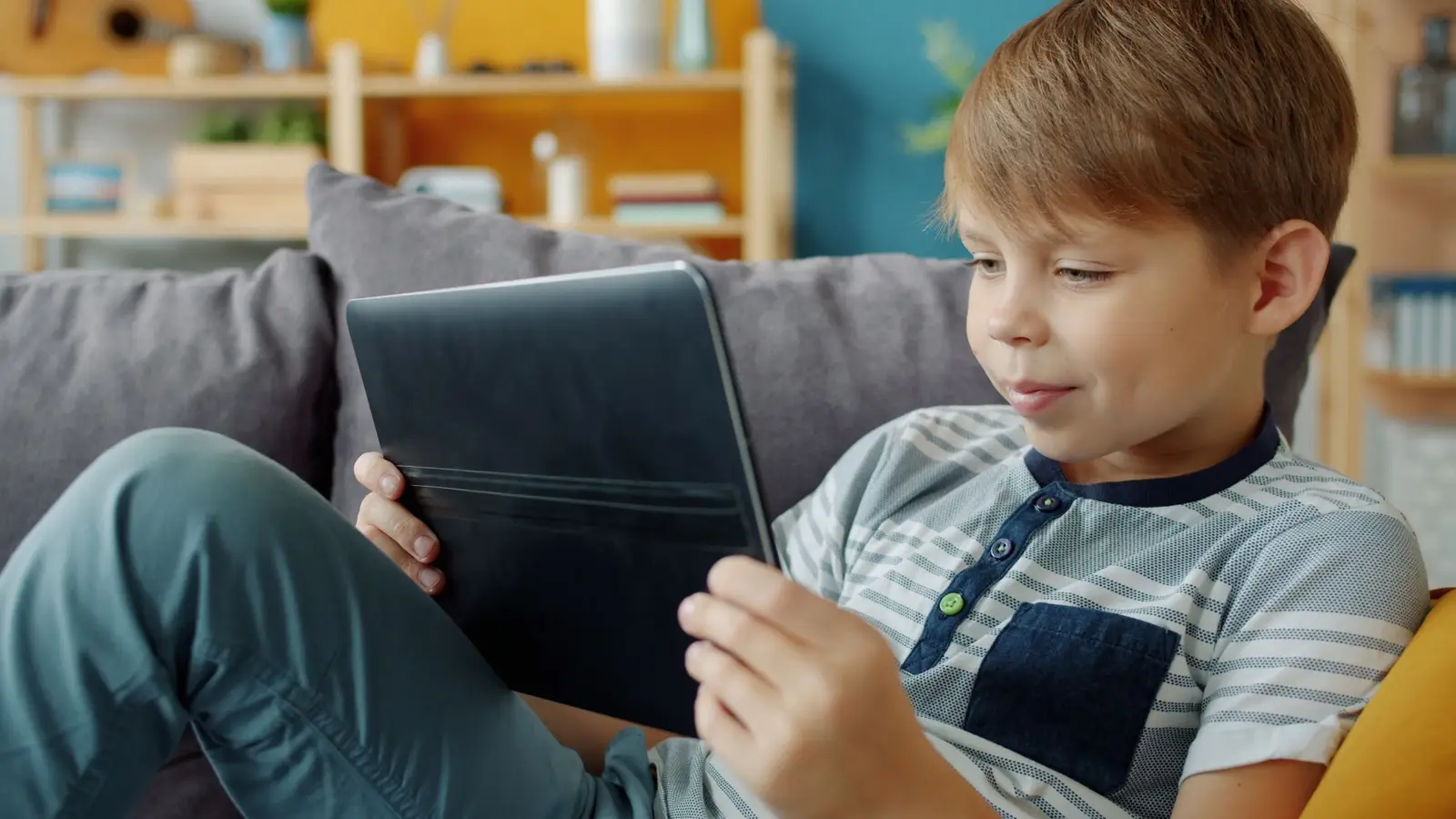 A boy smiling at a tablet while sitting on a sofa at home