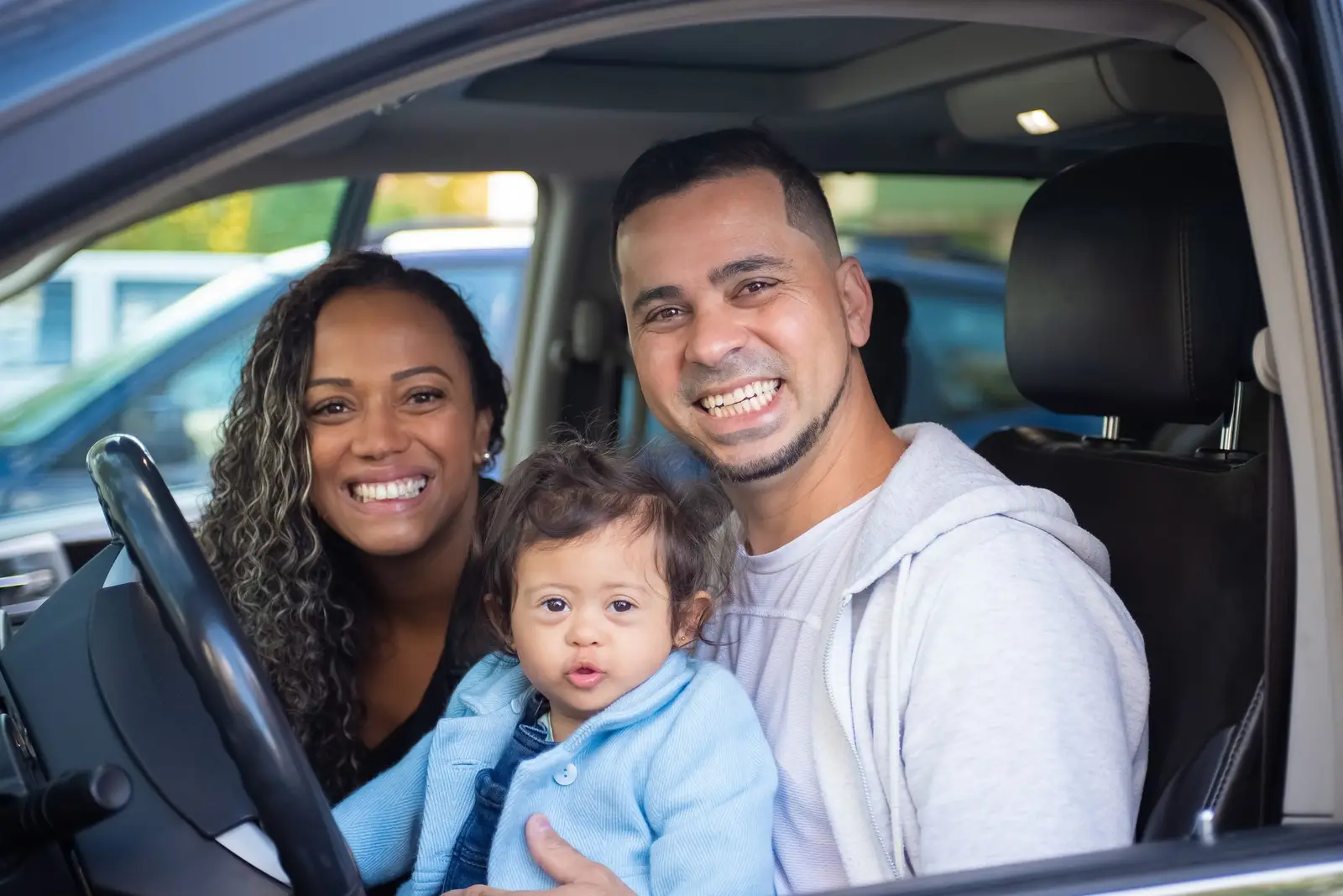 A smiling family with a baby sitting in the front of a car