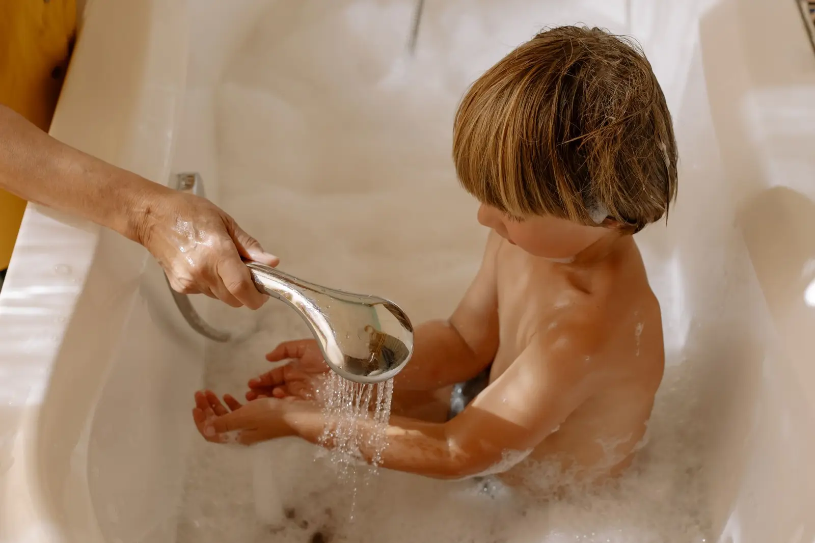 A young boy in a bubble bath while a parent rinses his hands with the shower head