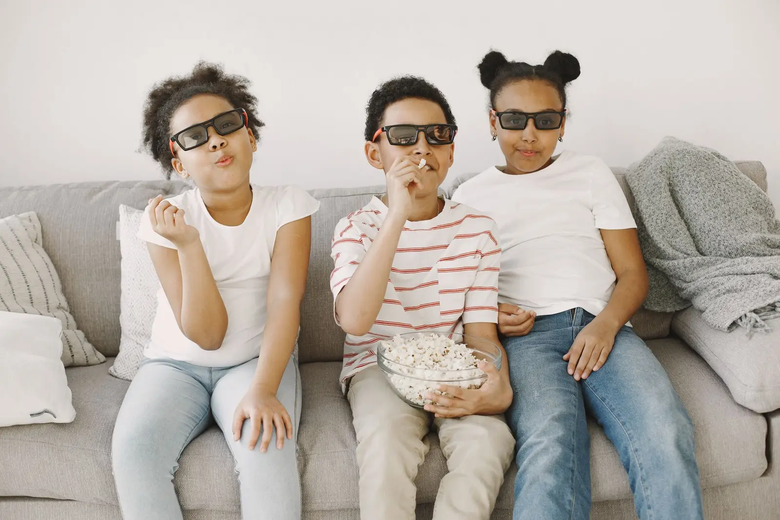 Three children in 3D glasses eating popcorn on a sofa