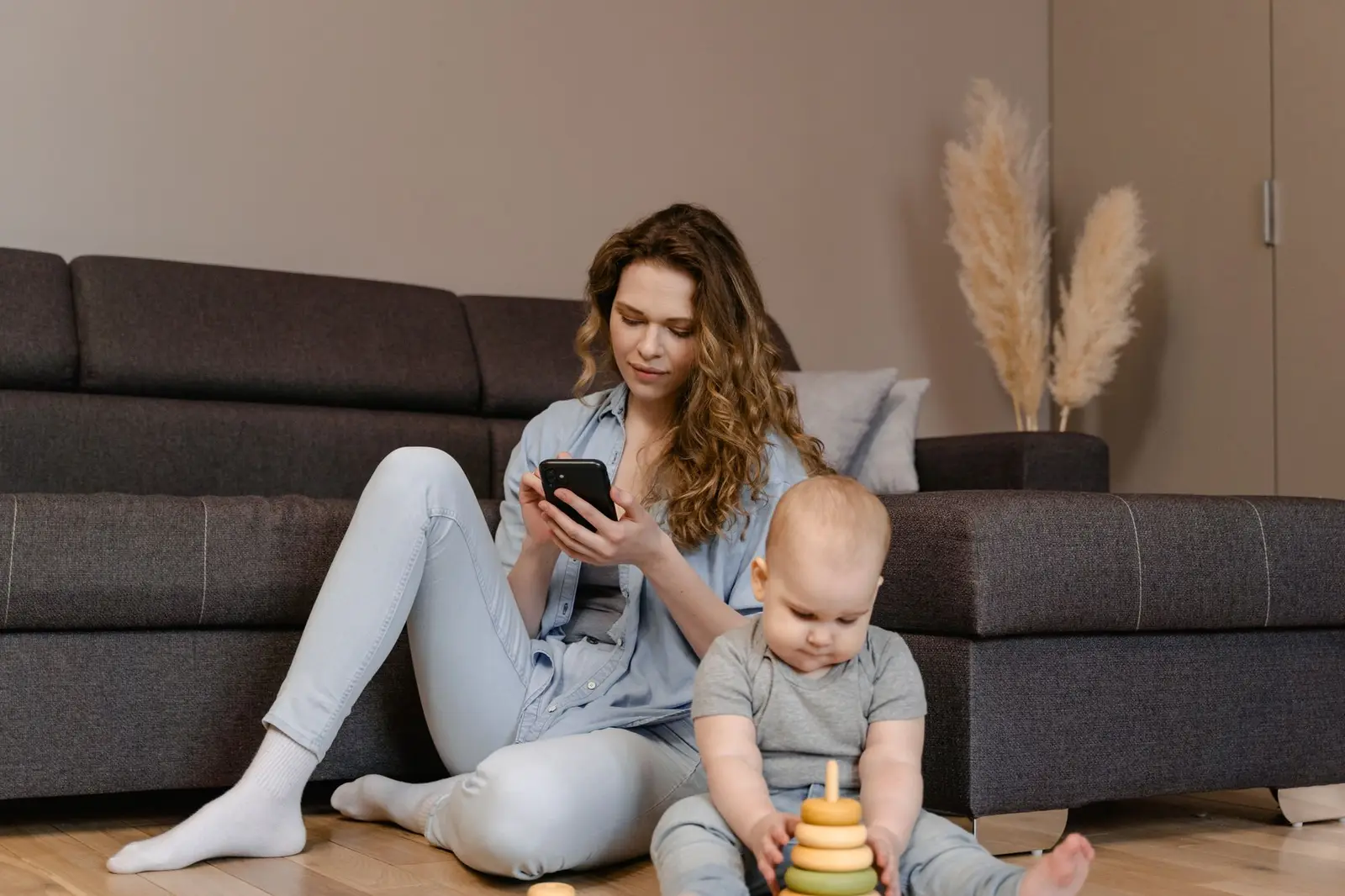 A mother on the living room floor looking at her phone while her baby plays with a stacking toy