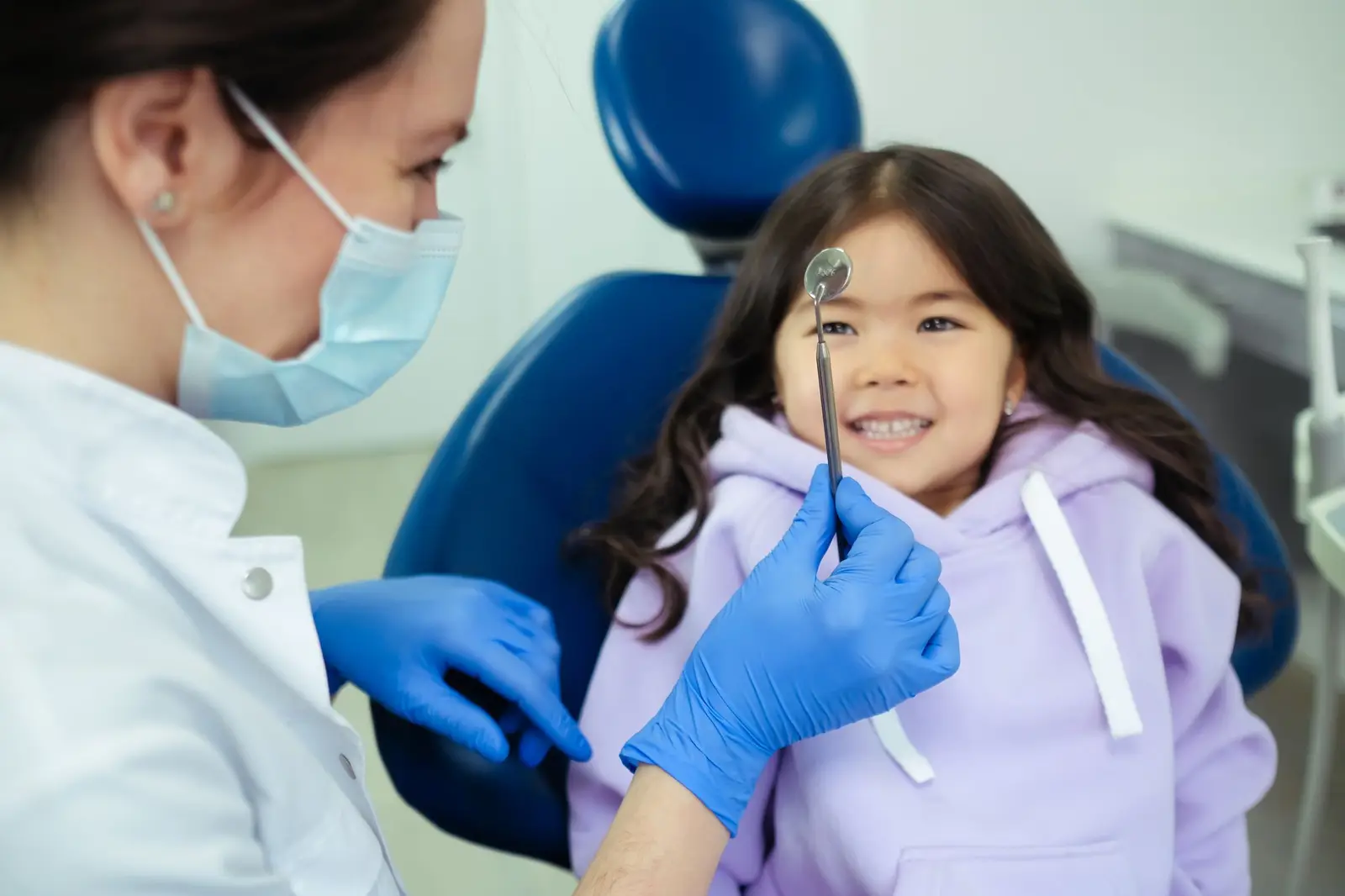 A smiling girl in a dental chair while a dentist holds a mirror