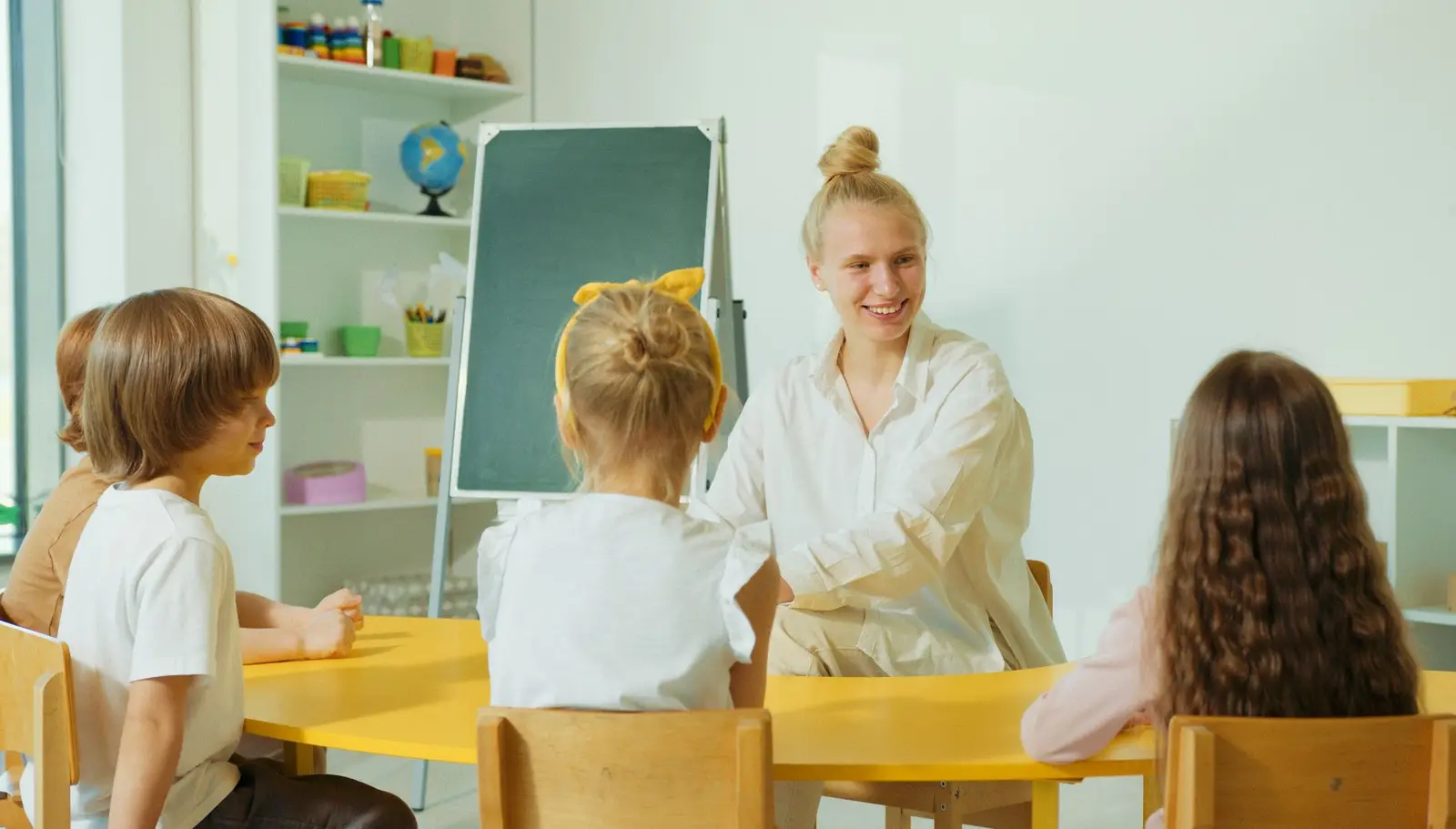 A teacher smiling at young children sitting at a table in a bright classroom