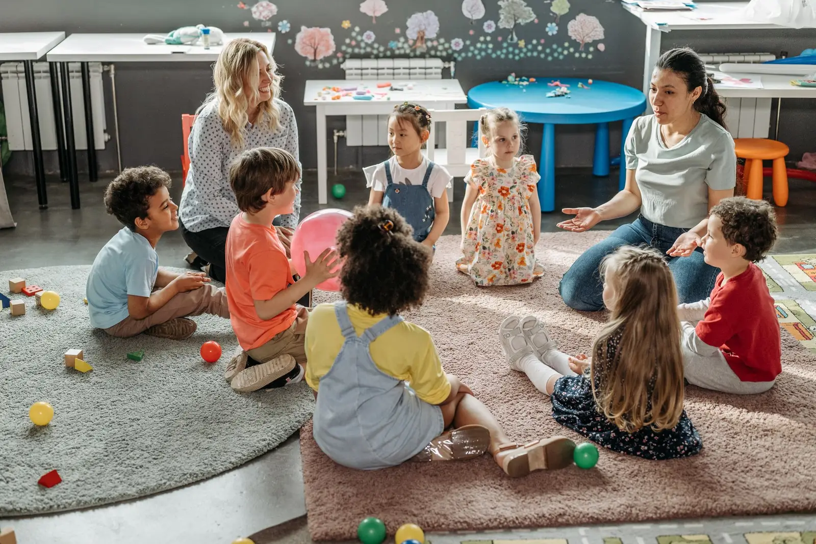 Young children sitting in a circle on a rug with two educators in a bright classroom