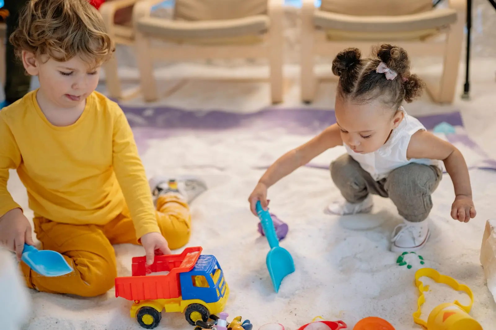 Two young children playing with toy trucks and spades in a sandpit