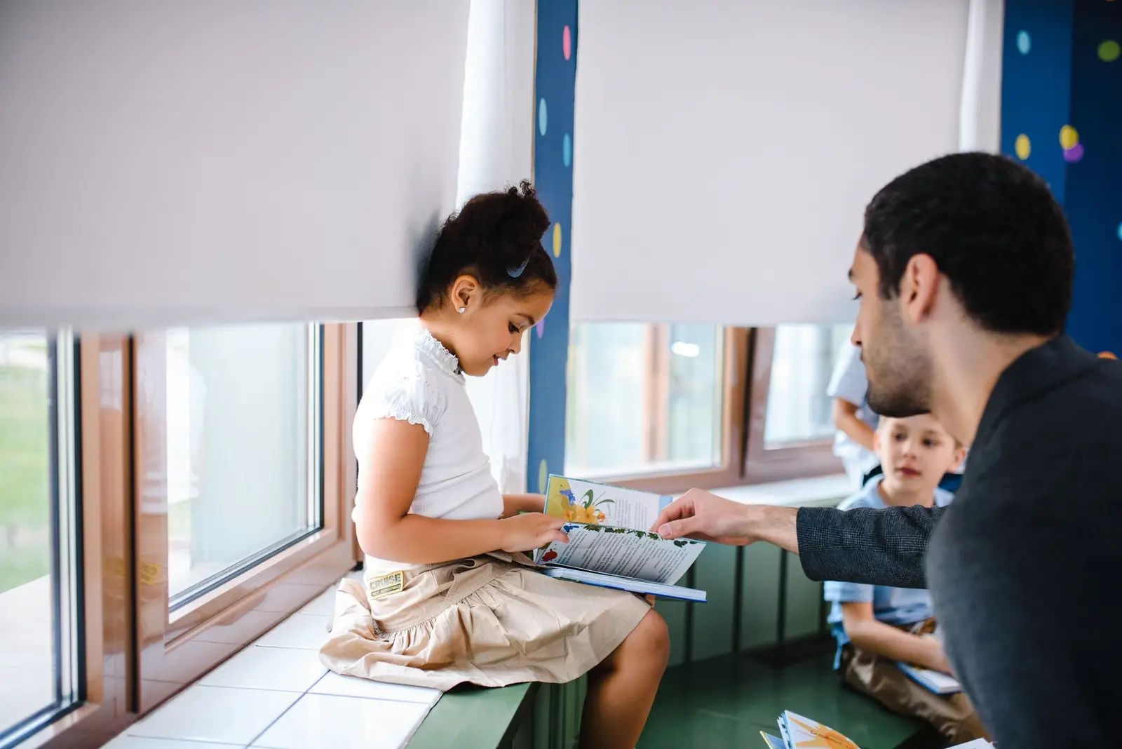 A teacher helping a young girl read a book by the classroom window