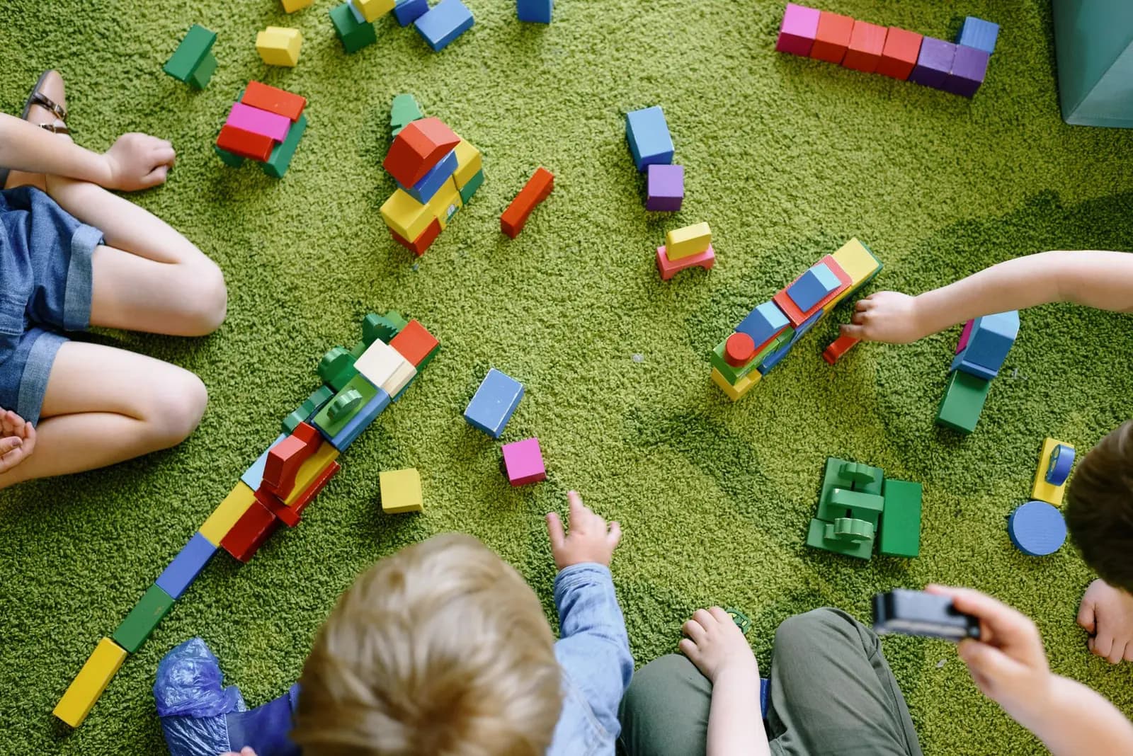 Children's hands playing with colourful wooden blocks on a green rug, seen from above